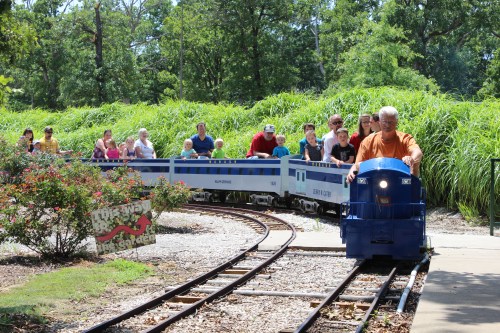 A miniature train with passengers travels along a track surrounded by greenery in a park setting.