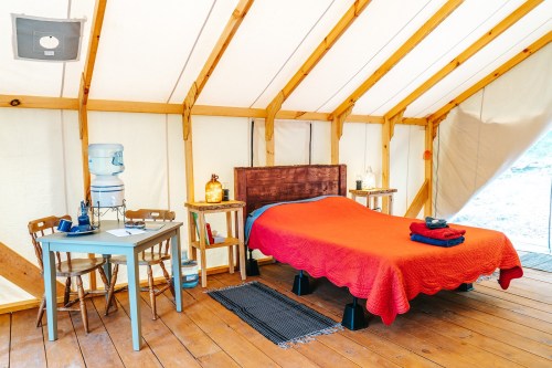 Cozy glamping tent interior with a red bedspread, wooden furniture, and a water cooler, set in a rustic environment.