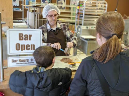 A woman in a hairnet prepares treats at a counter while a child and an adult watch. A sign indicates the bar is open.