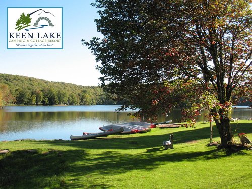 A serene lake view with canoes, green grass, and trees, featuring the Keen Lake Camping & Cottage Resort logo.