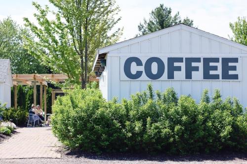 A white building with a large "COFFEE" sign, surrounded by greenery and outdoor seating in a sunny setting.