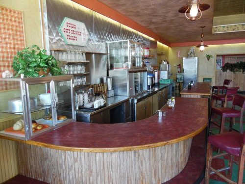 A retro diner interior featuring a curved counter, stools, and a stainless steel kitchen area with glass displays.