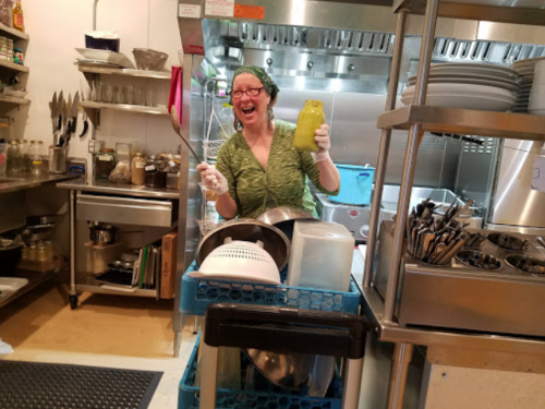 A smiling person in a green outfit holds a yellow jug in a busy kitchen, surrounded by pots and utensils.