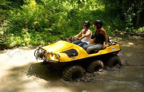 Two women ride a yellow all-terrain vehicle through muddy water in a lush green forest. Both wear helmets.