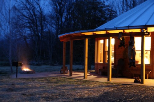 A cozy cabin at dusk, with warm light glowing from the windows and a fire pit nearby, surrounded by trees.