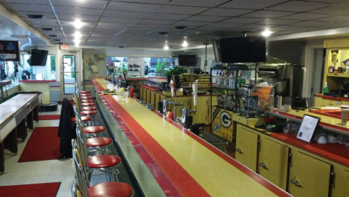 A retro diner interior featuring a long bar with red stools, colorful counters, and a variety of drinks on display.