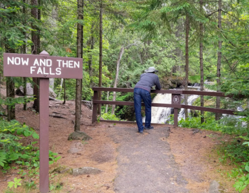A person leans over a wooden railing at Now and Then Falls, surrounded by lush green trees and a flowing waterfall.