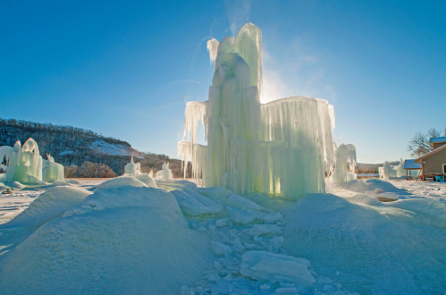 A frozen ice sculpture glistens in the sunlight, surrounded by snow and icy formations against a clear blue sky.