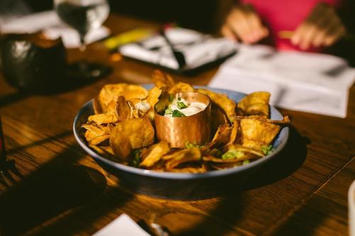 A blue bowl filled with crispy chips and a small copper cup of dip, garnished with green onions on a wooden table.