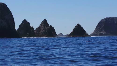 Rocky peaks rise from a deep blue ocean under a clear sky, creating a dramatic coastal landscape.