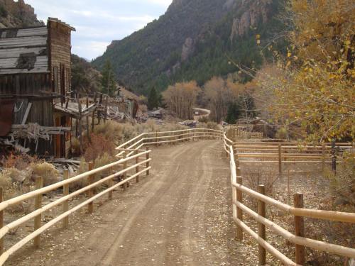 A dirt path winds through a rustic landscape with wooden fences, surrounded by mountains and autumn foliage.