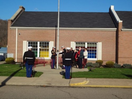 Marines in uniform salute as a group gathers outside a brick building with colorful windows, under a clear blue sky.