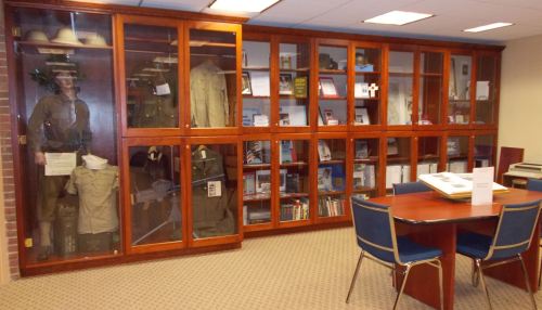Display case with military uniforms and memorabilia, alongside a table and chairs in a well-lit room.