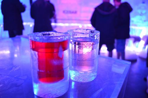 Two ice glasses filled with red and clear liquid, set against a colorful, illuminated background with people in the distance.