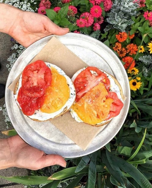 A silver plate holds two bagels topped with cream cheese and slices of red and yellow tomatoes, surrounded by colorful flowers.