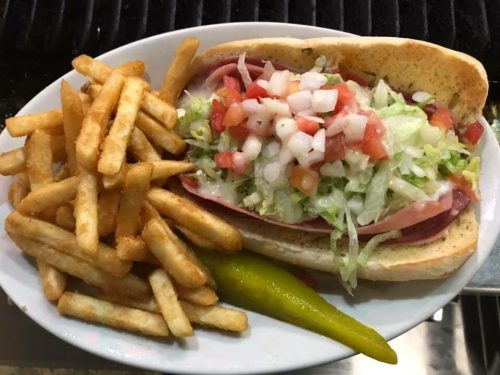 A sub sandwich filled with lettuce, tomatoes, and meats, served with fries and a pickle on the side.
