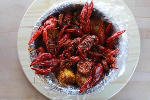 A bowl filled with spicy crawfish, corn, and sausage, all covered in seasoning and served on a wooden surface.
