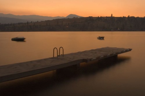 A serene lake at sunset, featuring a wooden dock and boats gently floating on calm waters.