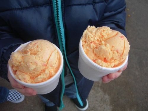 A person holds two cups of orange ice cream, wearing a blue jacket and standing on a concrete surface.