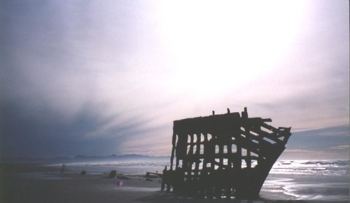 Silhouette of a shipwreck on a beach against a dramatic sky with soft clouds and distant mountains.