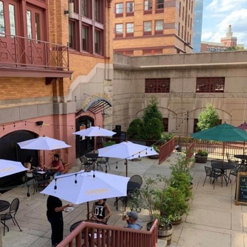 Outdoor café area with umbrellas, tables, and greenery in an urban setting. People are seated and enjoying the space.