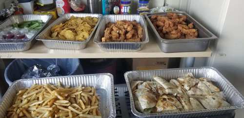 A table with various food trays: celery, chips, fried chicken, fries, and quesadillas in aluminum containers.