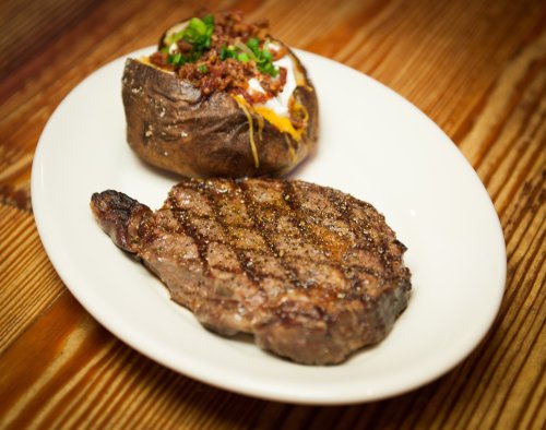 A grilled steak with a perfectly seared crust, served alongside a loaded baked potato topped with cheese and green onions.