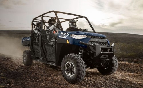 A blue off-road vehicle with three riders navigating a rugged terrain under a cloudy sky. Dust is kicked up behind it.