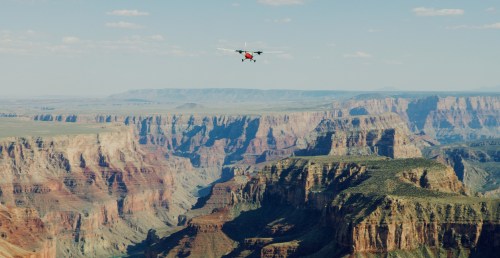 A drone flies over a vast canyon landscape with layered rock formations and distant mountains under a blue sky.