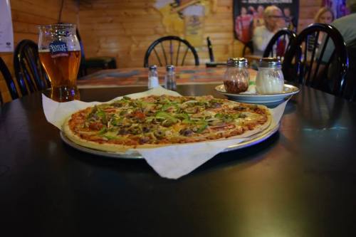 A large pizza topped with vegetables sits on a table next to a glass of beer and condiments in a cozy restaurant.