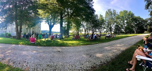 A group of people sitting on lawn chairs under trees, enjoying an outdoor event near a house and a gravel path.