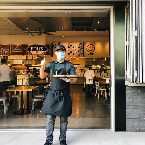 A masked server holds a plate outside a pizzeria, with a modern restaurant interior visible in the background.