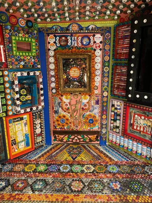 Colorful, intricate wall and floor design made of bottle caps, featuring a decorative mirror and various artistic elements.