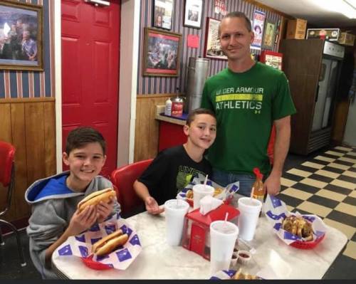 A man and two boys enjoy hot dogs and drinks at a diner with a retro decor.