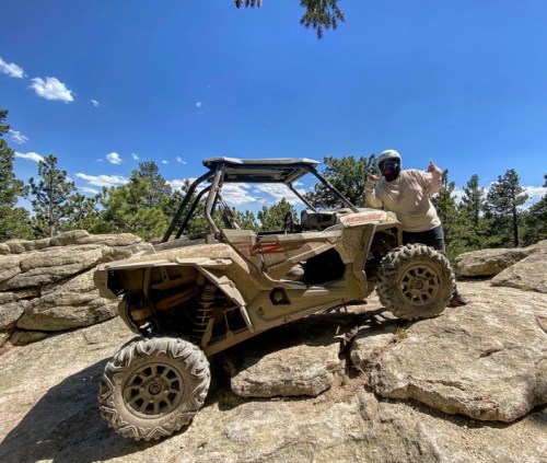 A person gives a thumbs up next to a muddy off-road vehicle on rocky terrain under a blue sky with clouds.