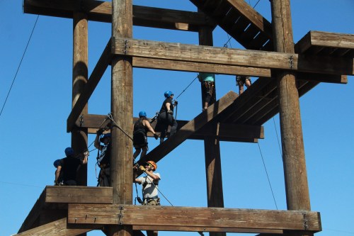 A group of people climbing a wooden structure with safety gear on a clear blue sky day.