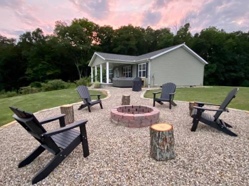 A cozy outdoor seating area with black chairs around a fire pit, set against a house and a colorful sunset sky.