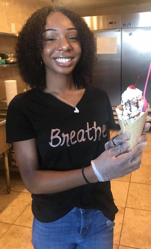 A smiling young woman holds an ice cream cone topped with chocolate syrup and a cherry, wearing a "Breathe" shirt.