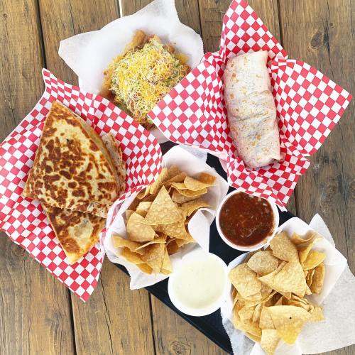A spread of Mexican food featuring a quesadilla, burrito, nachos with salsa, and cheese dip in red-checkered baskets.
