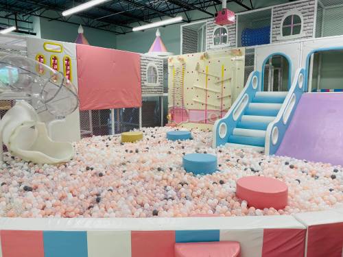 A colorful indoor play area featuring slides, climbing walls, and a ball pit filled with pastel balls.