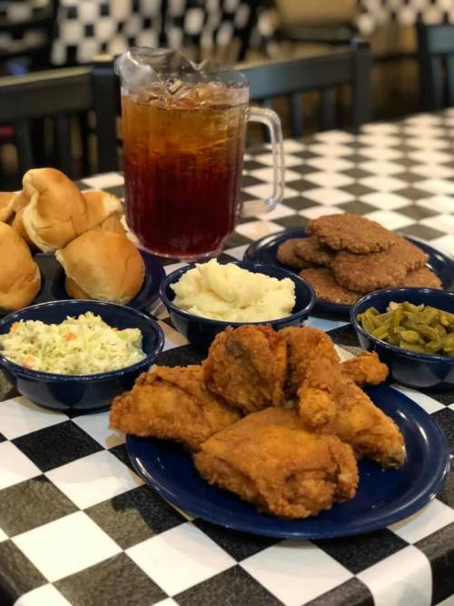 A spread of fried chicken, mashed potatoes, coleslaw, green beans, rolls, and iced tea on a checkered tablecloth.