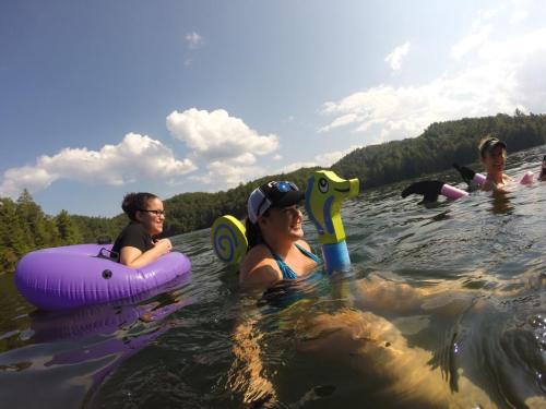 Three people float in a lake on inflatable toys, surrounded by trees and a blue sky with clouds.