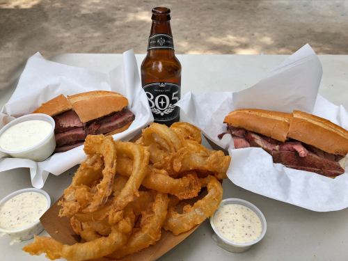 Two sandwiches with sliced meat, a serving of onion rings, and a beer bottle on a table. Dipping sauces on the side.