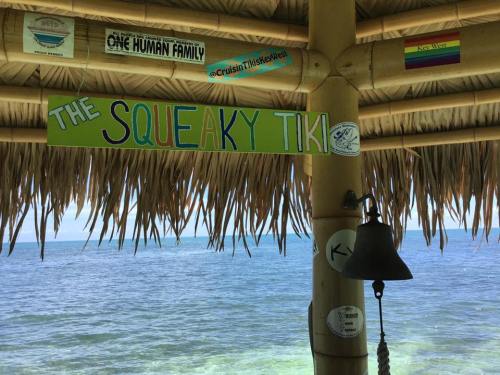 Sign reading "The Squeaky Tiki" under a thatched roof, overlooking a calm sea with various stickers nearby.