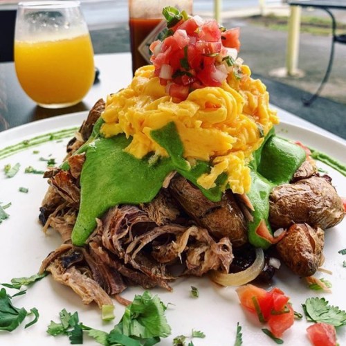 A colorful plate featuring shredded meat topped with mashed potatoes, green sauce, and fresh salsa, with a drink in the background.