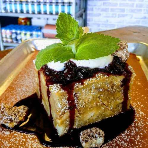 A square dessert topped with cream, mint, and berry sauce, served on a metal tray with a sprinkle of powdered sugar.