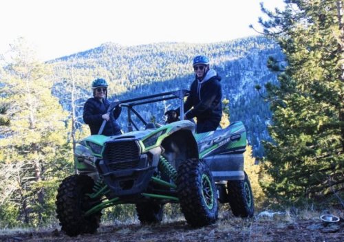 Two people wearing helmets sit on a green and black ATV in a forested area with mountains in the background.