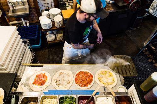 A chef prepares pizzas with various toppings in a bustling kitchen, steam rising from the cooking area.