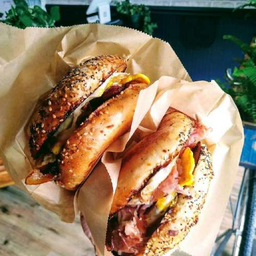 Two sesame seed bagels filled with various toppings, held in brown paper, against a backdrop of greenery.