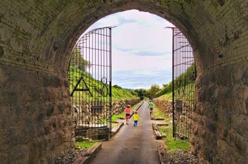 Two children walk down a path through a stone archway, flanked by green hills and a cloudy sky.